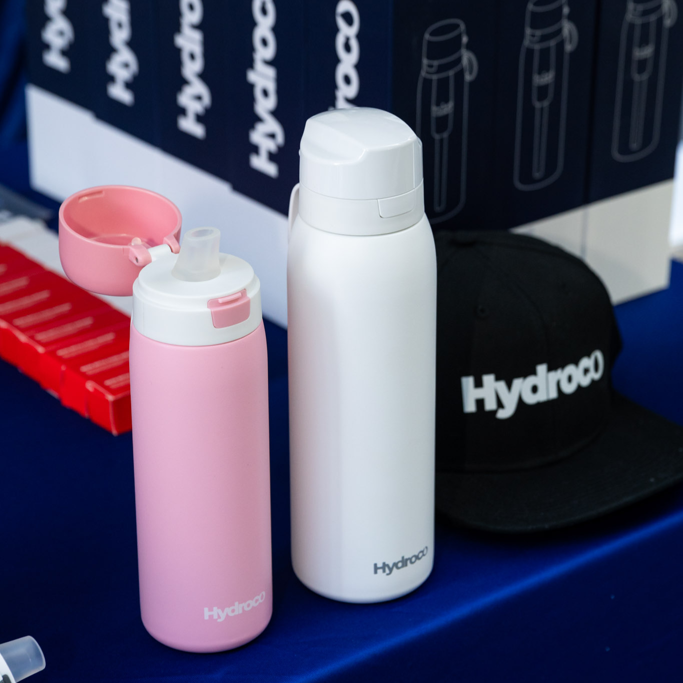 Two Hydroco water bottles, one pink and one white, on a blue surface with boxes in the background.