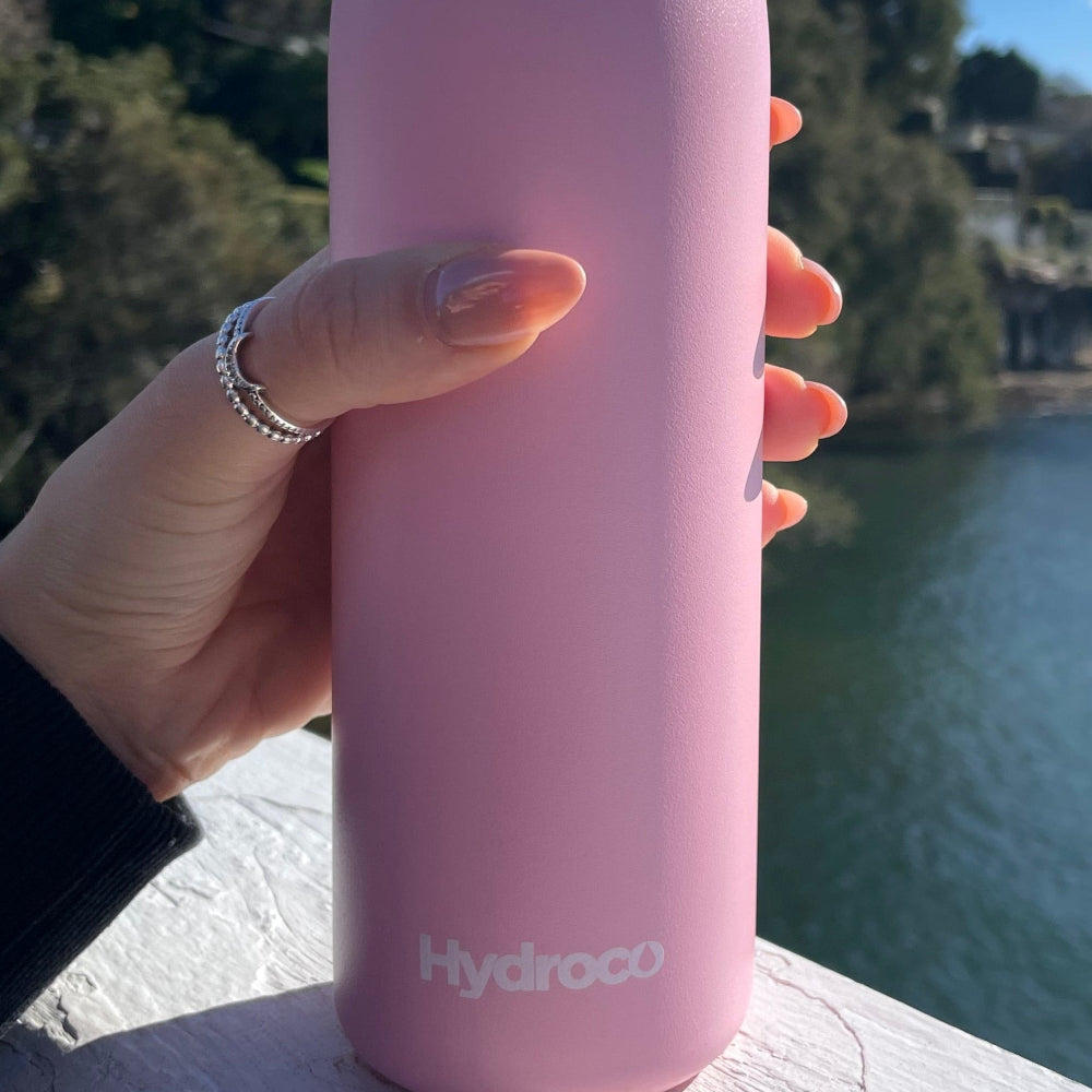 Hand holding a pink Hydroco Flask against a scenic background with water and sky.